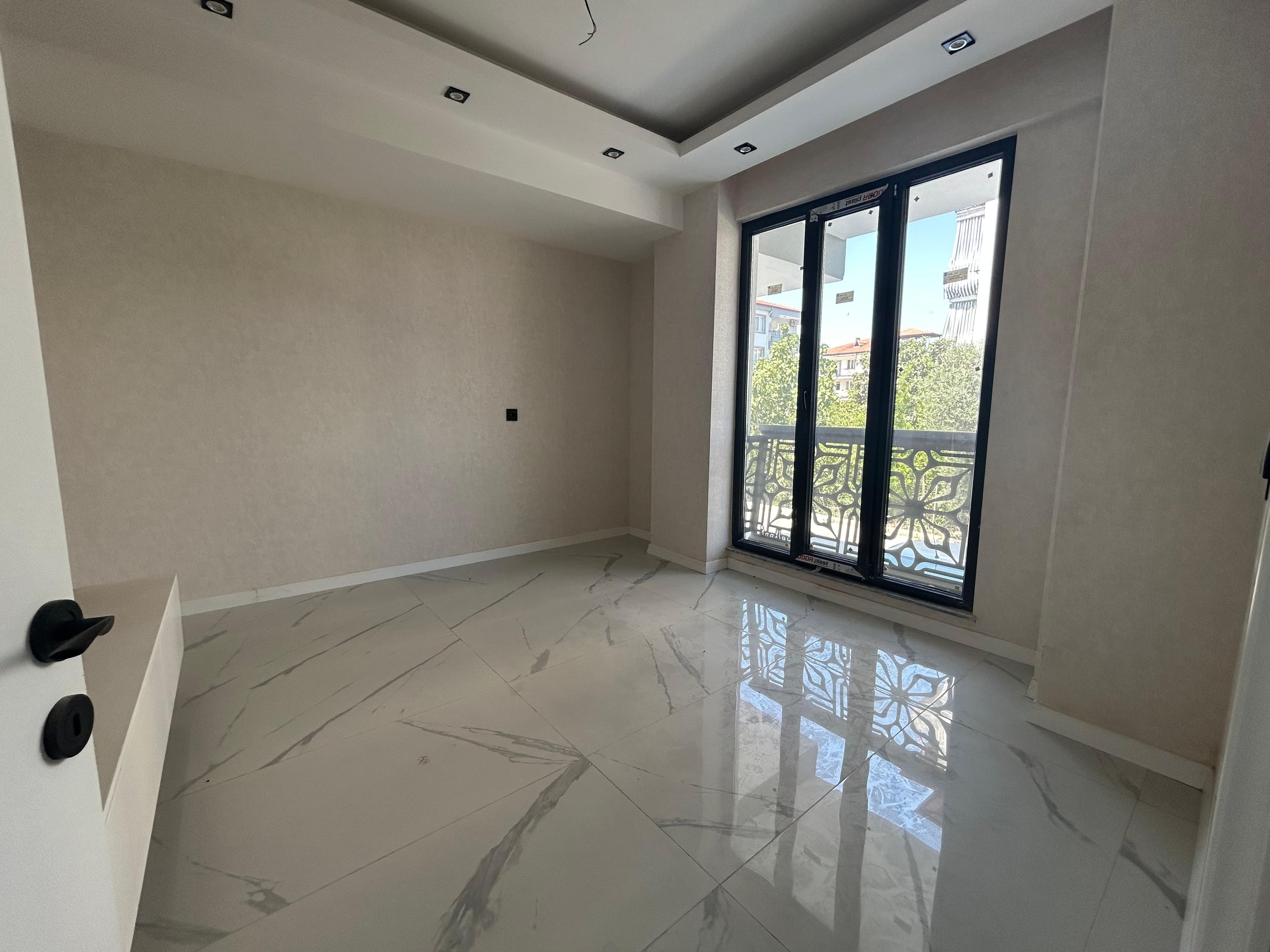 Boş, yenilenmiş bir oda; mermer desenli zemin, kademeli alçıpan tavan spot aydınlatmalı, siyah çerçeveli balkon kapısı ve dövme demir korkuluk.