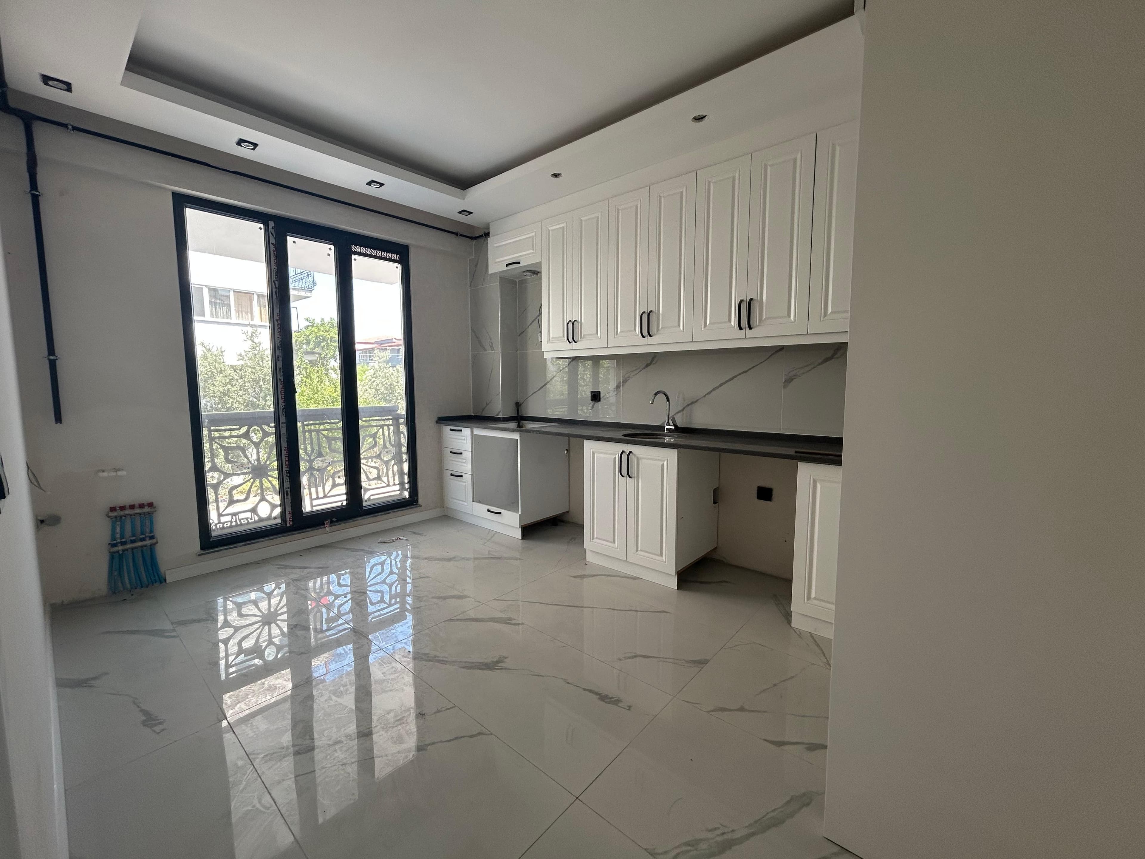 Açık mutfak-salon; beyaz kabartma kapaklı üst-alt dolaplar, siyah tezgah, mermer desenli sırt paneli, mermer desenli zemin, balkon kapısı.