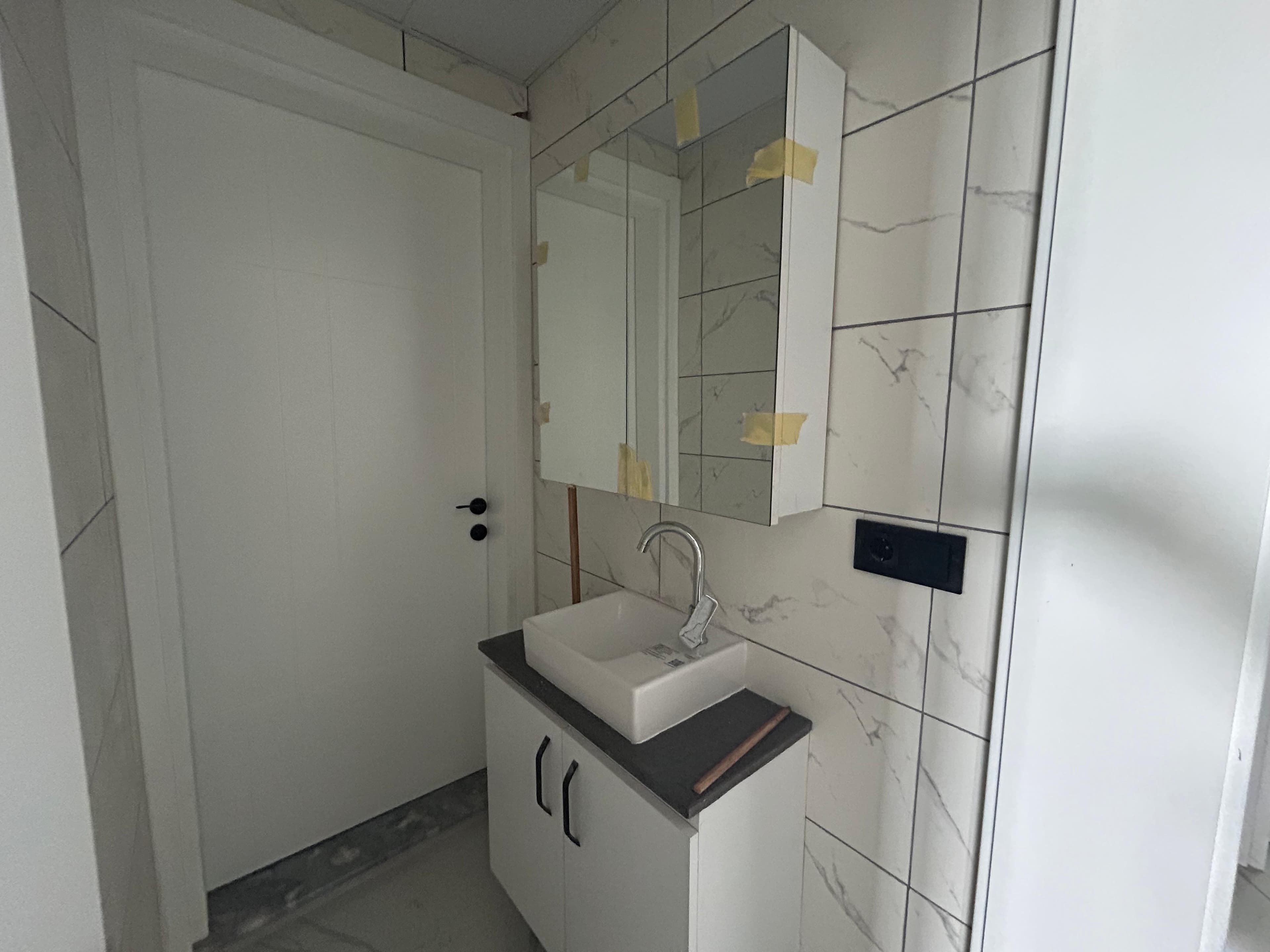 Kurulum aşamasında banyo; koruma bantlı aynalı dolap, tezgah üstü lavabo, mermer desenli fayans, beyaz iç kapı.
