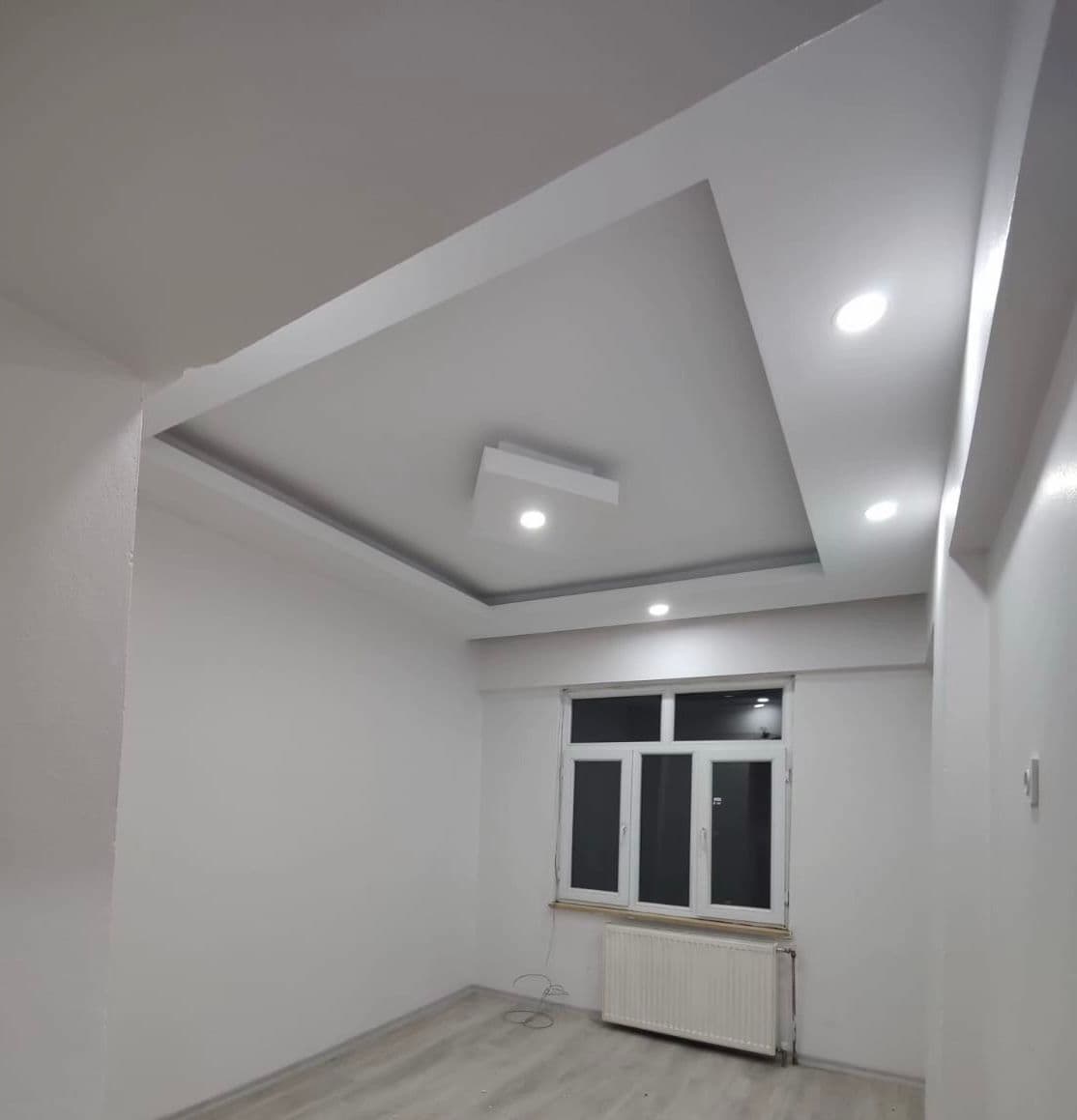 Bitmiş kademeli alçıpan tavan köşe detayı; spot aydınlatmalar, pencere ve petek radyatör, laminat zemin.